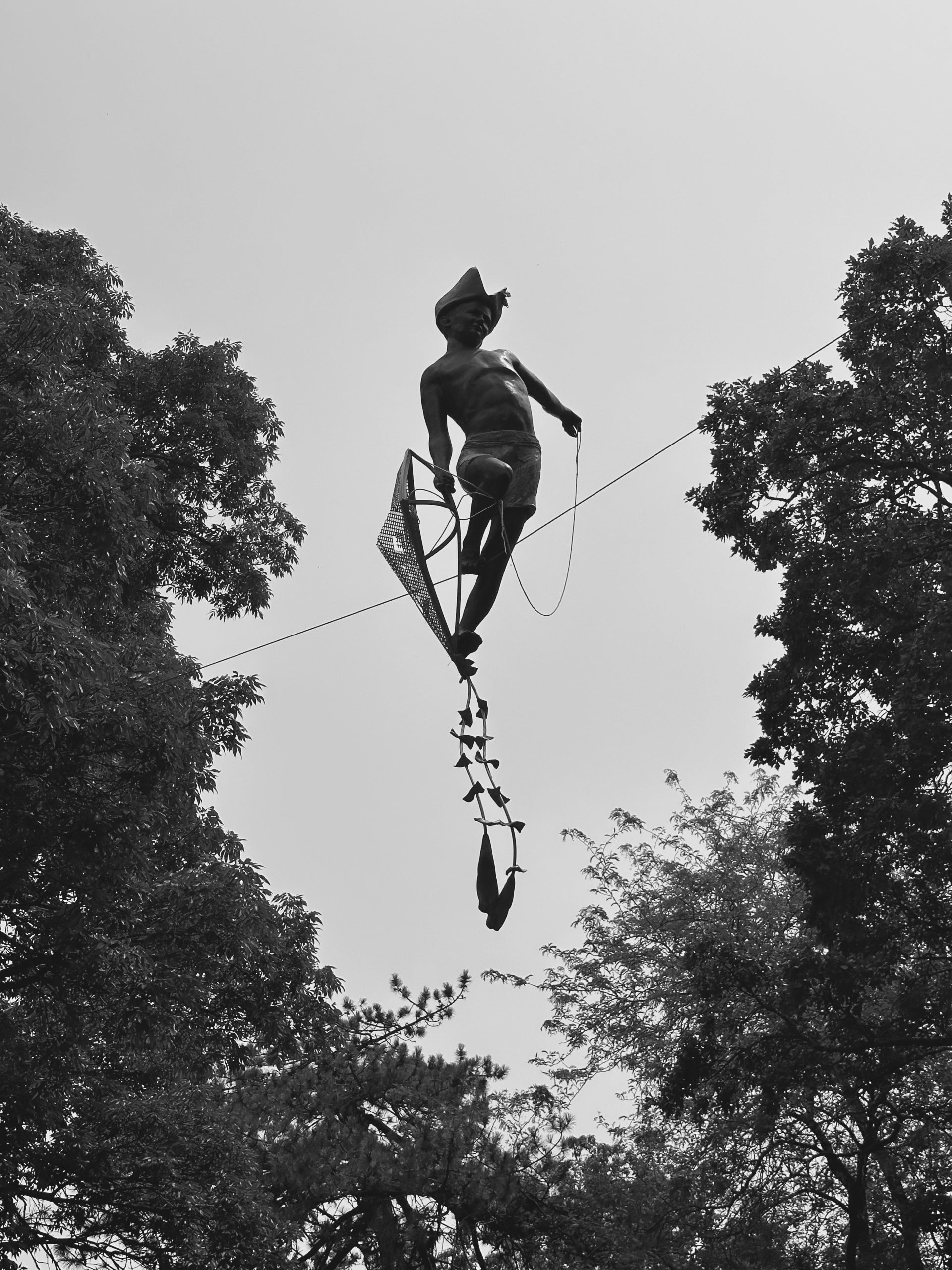 A statue of a boy holding a kite and wearing a hat is suspended in the air between two trees.